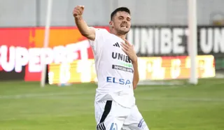 Un bărbat cu tricou alb și pantaloni albi de sport se află pe un teren de fotbal verde, având un braț ridicat.