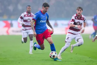 Un jucător într-un echipament albastru și roșu conduce un balon pe un teren de fotbal, fiind urmărit de alți doi jucători.