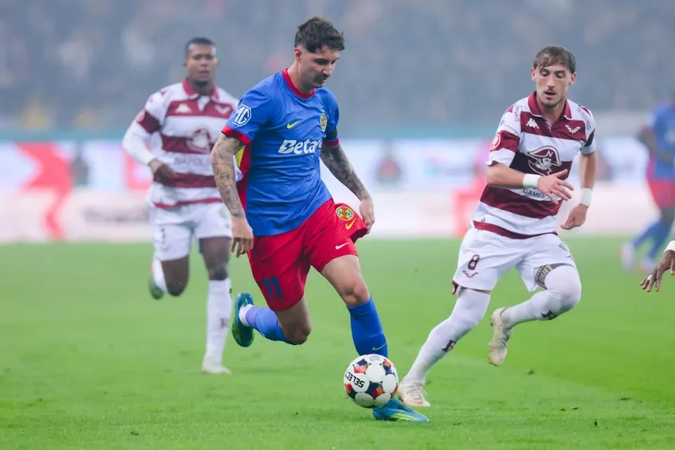 Un jucător într-un echipament albastru și roșu conduce un balon pe un teren de fotbal, fiind urmărit de alți doi jucători.