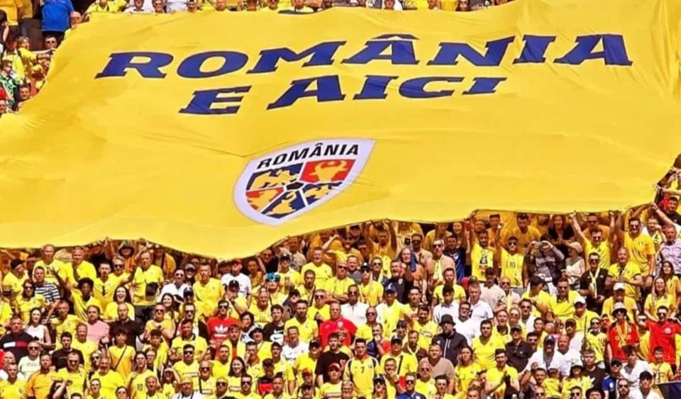O mulțime de oameni îmbrăcați în tricouri galbene ridică un steag mare de culoare galbenă pe care scrie text albastru și un emblema colorată.