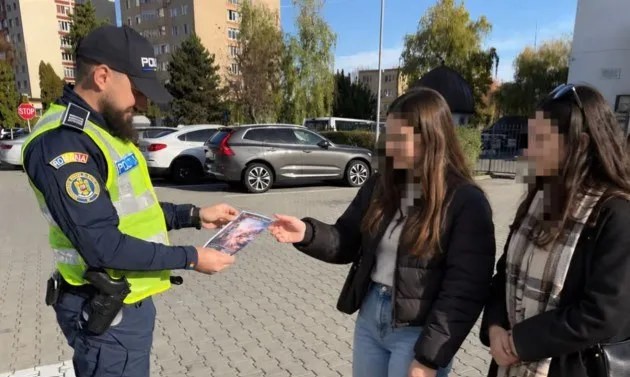 Un polițist în uniformă și vestă reflectorizantă oferă un document unei femei într-un spațiu exterior cu mașini și clădiri în fundal.