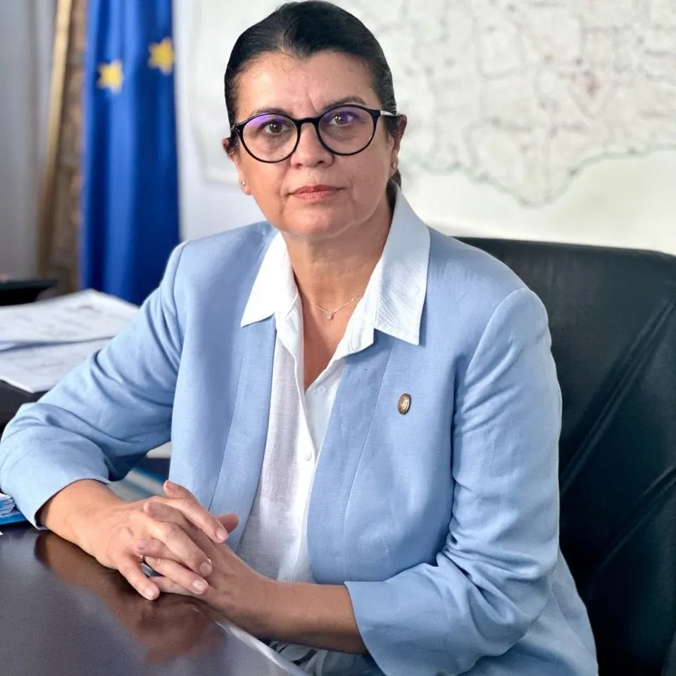 O femeie cu ochelari și sacou albastru stă la o masă cu mâinile împreunate în față.