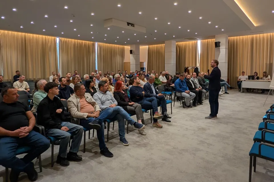 Un bărbat stă în picioare în fața unui grup de persoane așezate pe scaune într-o sală cu perdele lungi și lumină de plafon.