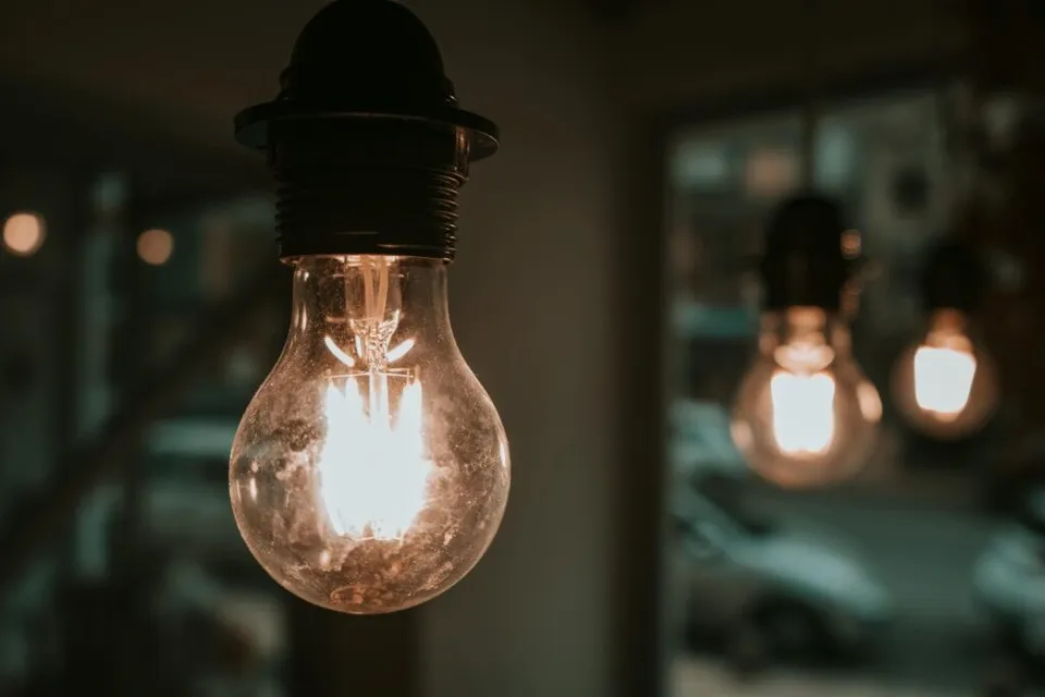 O bec incandescentă aprinsă este suspendată într-un decor întunecat, cu alte becuri luminoase, neclare, în fundal.