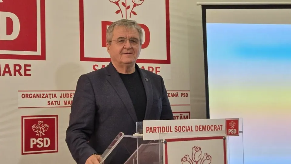 Un bărbat cu ochelari și sacou închis la culoare stă în fața unui podium cu logoul PSD și un panou informativ în spate.