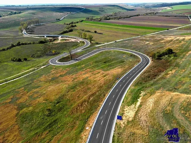 O fotografie aeriană prezintă o șosea asfaltată cu linii albe care serpuează printr-un peisaj de dealuri verzi și câmpuri agricole sub un cer deschis.