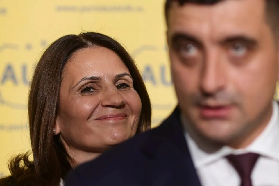 O femeie zâmbește în fundal, în timp ce un bărbat cu cămașă albă și cravată purpurie apare neclar în prim-plan.