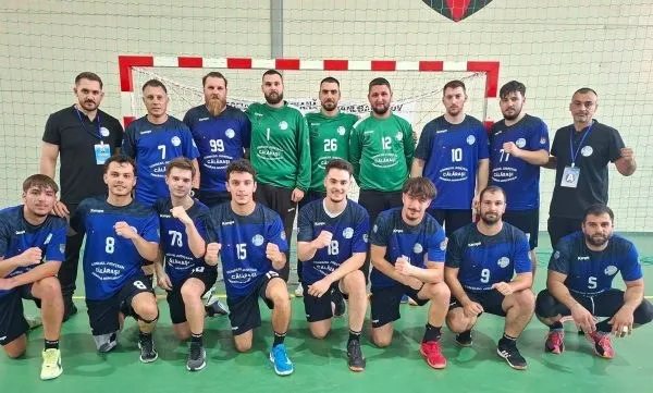 O echipă de handbal de unsprezece jucători și un antrenor pozează într-o sală de sport pe un teren verde.
