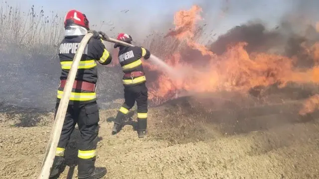 Doi pompieri în echipament de protecție negru cu benzi galbene stau cu spatele și folosesc un furtun pentru a stinge flăcările de pe un teren.