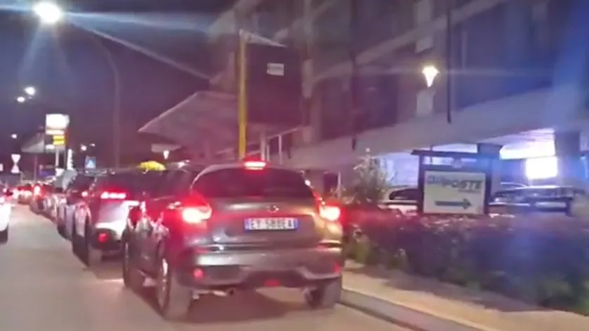O coadă de mașini stă la semafor sau într-o stație de alimentare pe timp de noapte, sub lumini artificiale.