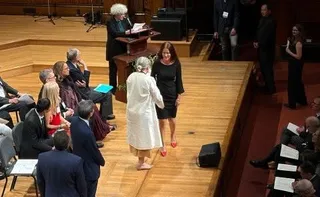O femeie în rochie neagră stă pe o scenă de lemn lângă o femeie în haine albe, în timp ce o altă persoană citește de la un pupitru.