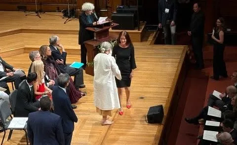 O femeie în rochie neagră stă pe o scenă de lemn lângă o femeie în haine albe, în timp ce o altă persoană citește de la un pupitru.