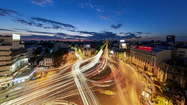 O vedere de sus asupra unui intersecție urbană iluminată noaptea, cu urme luminoase de la vehicule care traversează circulația.