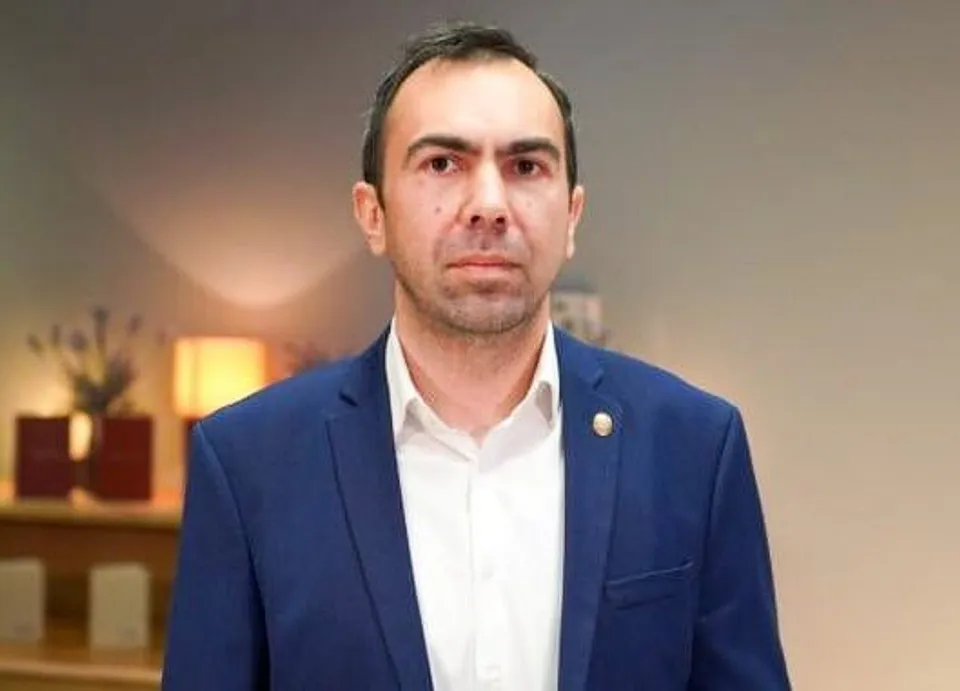 Un bărbat cu părul scurt și scurtă barbă poartă un sacou albastru peste o cămașă albă, stând în fața unui fundal neclar.