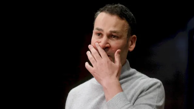 Un bărbat cu părul scurt și hanorac gri își ține mâna pe față în fața unui fundal întunecat.