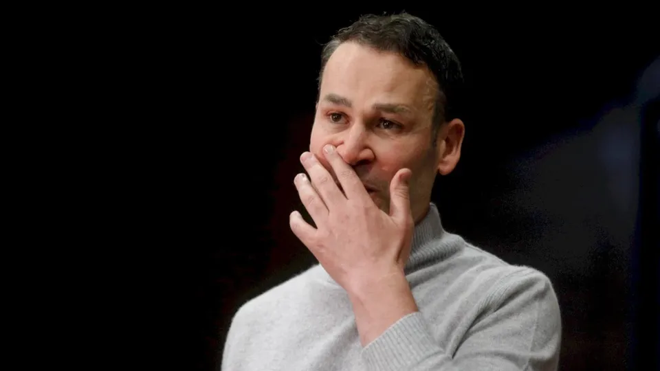 Un bărbat cu părul scurt și hanorac gri își ține mâna pe față în fața unui fundal întunecat.