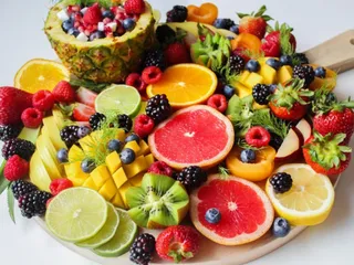 O tavă de lemn prezintă o varietate de fructe tăiate, inclusiv ananas, kiwi, citrice, căpșuni, murături și afine, dispuse pe un fundal alb.