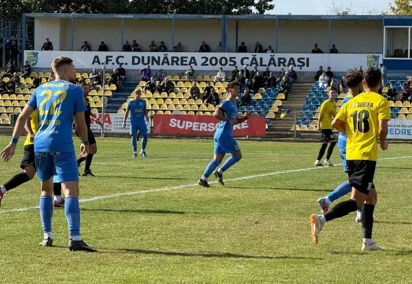 Câțiva jucători de fotbal în echipamente albastre și galbene aleargă pe un teren de gazon verde sub un cer senin.
