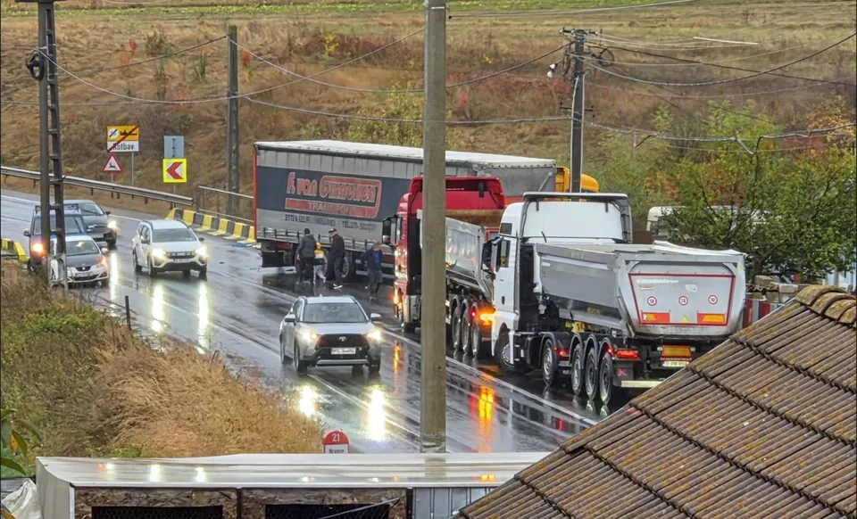 Un camion mare și un trailer sunt oprite pe un drum umed lângă un vehicul alb și alte mașini sub un cer înnorat.