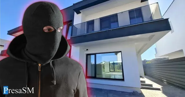 Un individ purtând o mască neagră de tip balaclava și o hanoracă neagră stă în prim-plan în fața unei clădiri albe cu balcon.