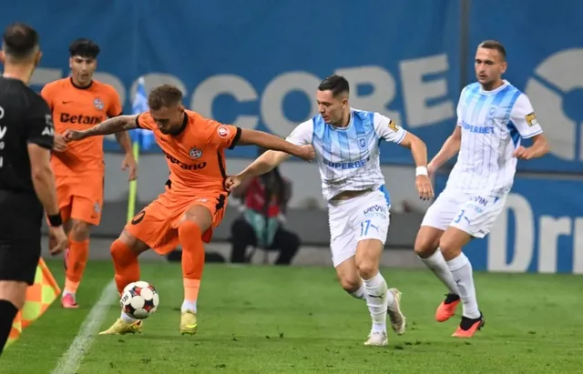 Un jucător în echipă portocalie lovește mingea pe un teren de fotbal în fața unui adversar în echipă albă și albastră.
