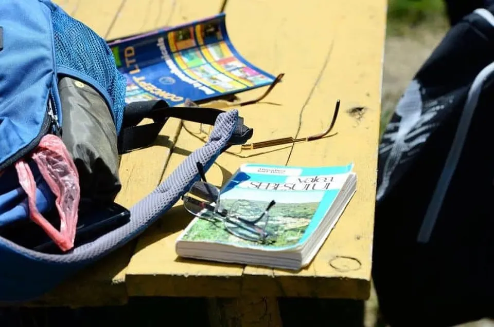 Un rucsac albastru, o carte cu ochelari de vedere și un alt obiect colorat sunt așezate pe o masă de lemn sub lumină solară.