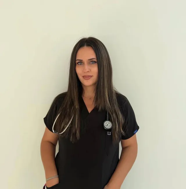 O femeie cu părul lung și deschis la culoare poartă un halat medical negru și un stetoscop în jurul gâtului, stând în fața unui perete alb.
