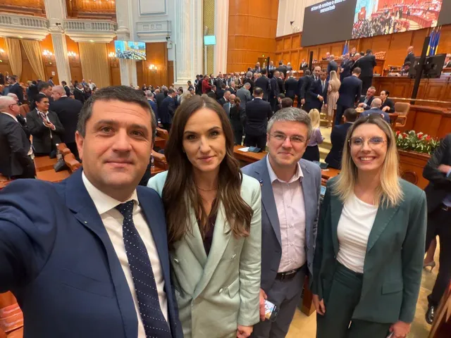 Patru persoane fac un selfie în prim-plan, în timp ce mulți oameni stau în scaune într-o sală de conferințe mare.