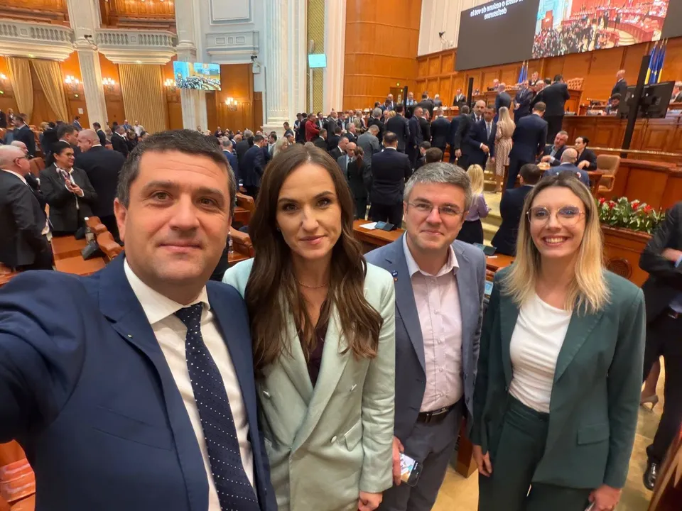 Patru persoane fac un selfie în prim-plan, în timp ce mulți oameni stau în scaune într-o sală de conferințe mare.