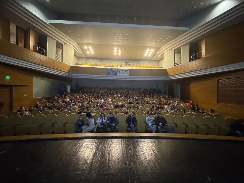 O mulțime de oameni stă așezată în scaunele unui cinema sau de la un eveniment într-o sală de spectacol cu pereți din lemn și tavan iluminat.