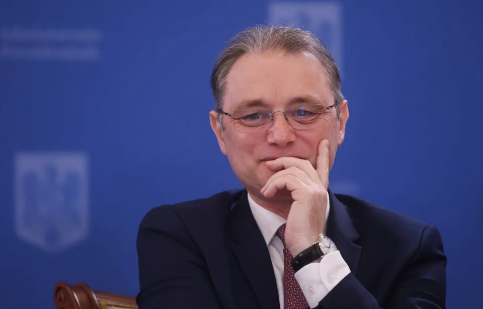 Un bărbat cu ochelari și costum închis la culoare stă cu mâna sub bărbie în fața unui fundal albastru.