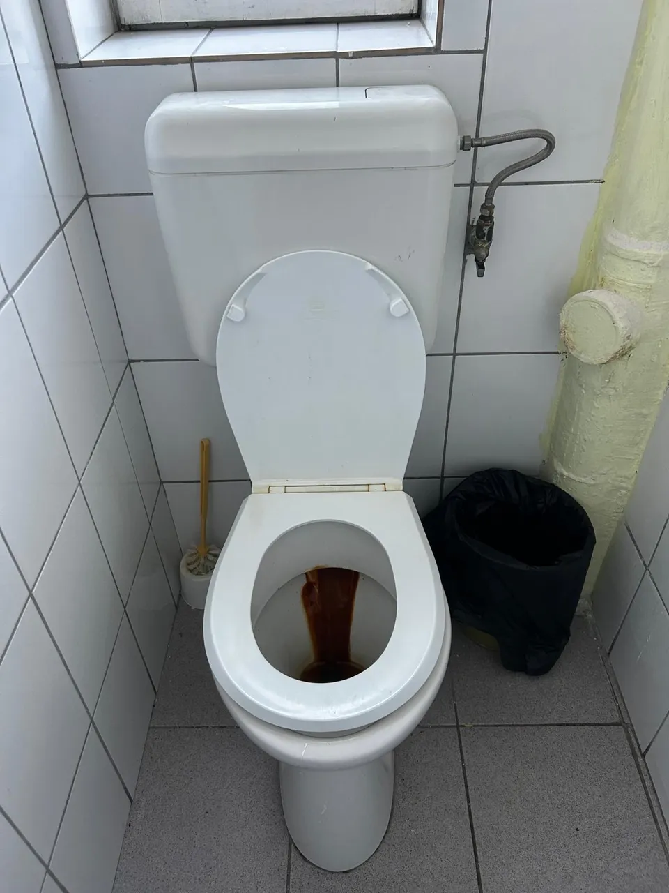 Un toaletă albă cu capacul ridicat este situată într-o încăpere cu plăci ceramice albe, lângă un coș de gunoi negru și o perie de curățare.
