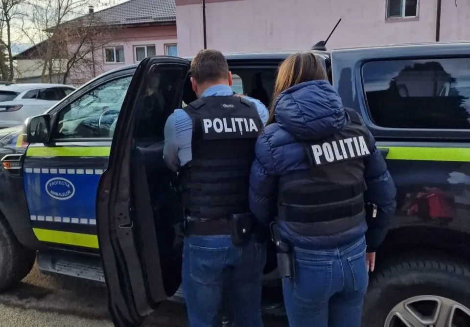 Doi polițiști în uniformă, cu veste negre pe care scrie POLIȚIA, stau lângă o mașină de serviciu neagră cu detalii galben-albastre.