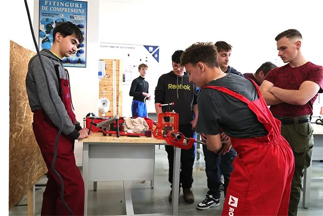 Un grup de tineri stă în jurul unei mese pe care se află unelte și piese mecanice într-un atelier.