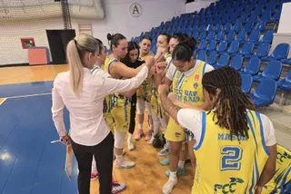 Un grup de femei îmbrăcate în echipamente sportive galbene și albe stau într-un cerc pe un teren de baschet lângă o persoană în cămașă albă.
