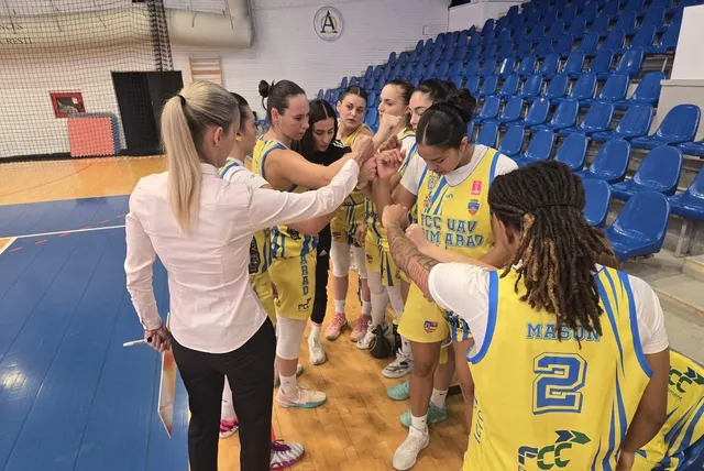 Un grup de femei îmbrăcate în echipamente sportive galbene și albe stau într-un cerc pe un teren de baschet lângă o persoană în cămașă albă.