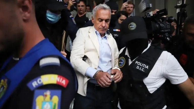 Un bărbat cu părul cărunt, purtând sacou deschis și cămașă albastră, merge printre persoane în uniformă și personal cu echipamente video.