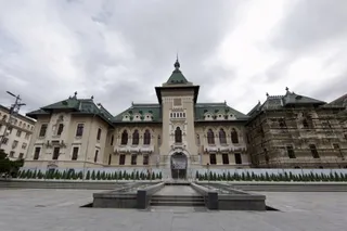 O clădire monumentală cu acoperiș verde și fațadă deschisă la culoare este situată în spatele unui spațiu pavat cu o fântână în prim-plan.