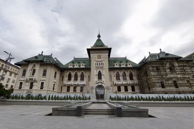 O clădire monumentală cu acoperiș verde și fațadă deschisă la culoare este situată în spatele unui spațiu pavat cu o fântână în prim-plan.