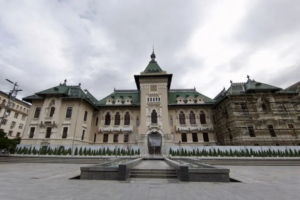 O clădire monumentală cu acoperiș verde și fațadă deschisă la culoare este situată în spatele unui spațiu pavat cu o fântână în prim-plan.