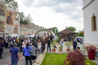 O mulțime de oameni stă într-o curte lângă o structură cu acoperiș transparent și o clădire cu un acoperiș în formă de piramidă.
