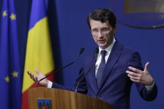 Un bărbat cu ochelari și costum în bleu portrivit vorbește de la un podium în fața unor steaguri.