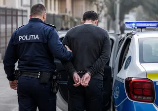 Un polițist în uniformă albastră stă lângă un bărbat cu mâinile în spate, în lanțuri, lângă o mașină de poliție albă și albastră.