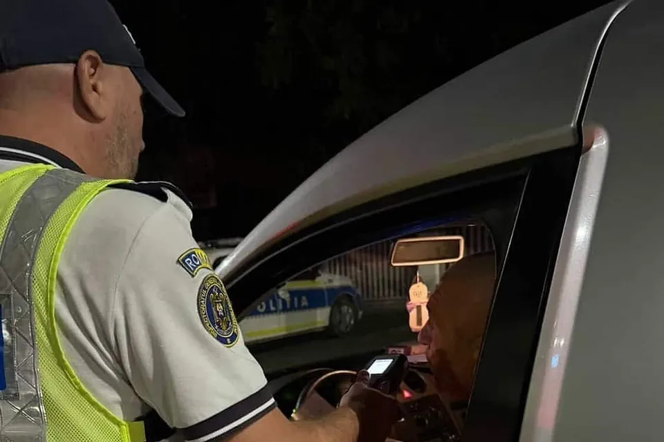 Un polițist în uniformă și vestă reflectorizantă verifică un document în interiorul unui autoturism alb pe timp de noapte.