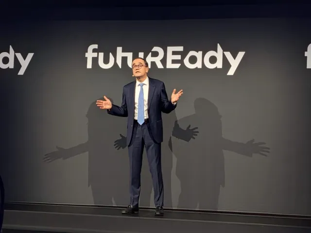 Un bărbat cu ochelari și cravată albastră stă pe o scenă în fața unui fundal gri cu textul white 'fuhuREady'.