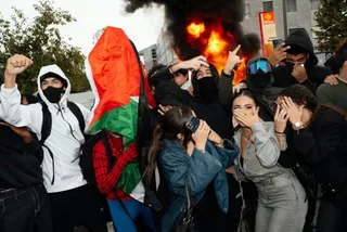 Un grup de tineri stă într-o mulțime lângă un nor de fum și flăcări, unii acoperindu-și fețele cu mâinile.