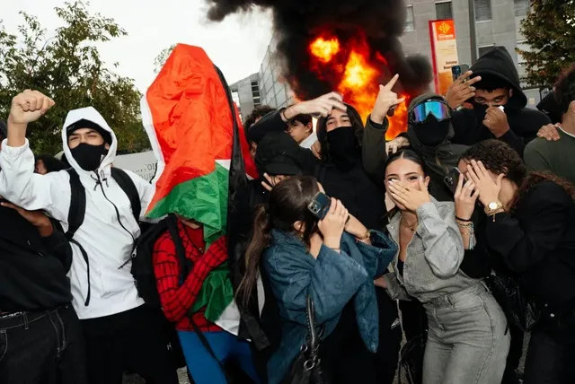 Un grup de tineri stă într-o mulțime lângă un nor de fum și flăcări, unii acoperindu-și fețele cu mâinile.