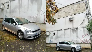 Un autoturism gri este parcat lângă un perete alb cu graffiti, acoperit de resturi de tencuială și frunze căzute pe sol.