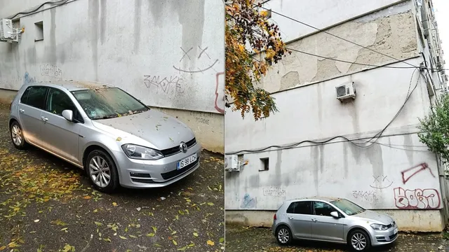 Un autoturism gri este parcat lângă un perete alb cu graffiti, acoperit de resturi de tencuială și frunze căzute pe sol.