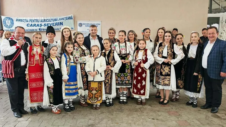 Un grup de persoane, inclusiv copii, poartă costume tradiționale albe cu broderii colorate și stau în fața unui perete deschis lângă un banner albastru.